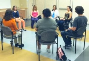 Group of adult men and women sitting in chairs in a circle with feet on the floor, hands in their lap, and eyes closed meditating.